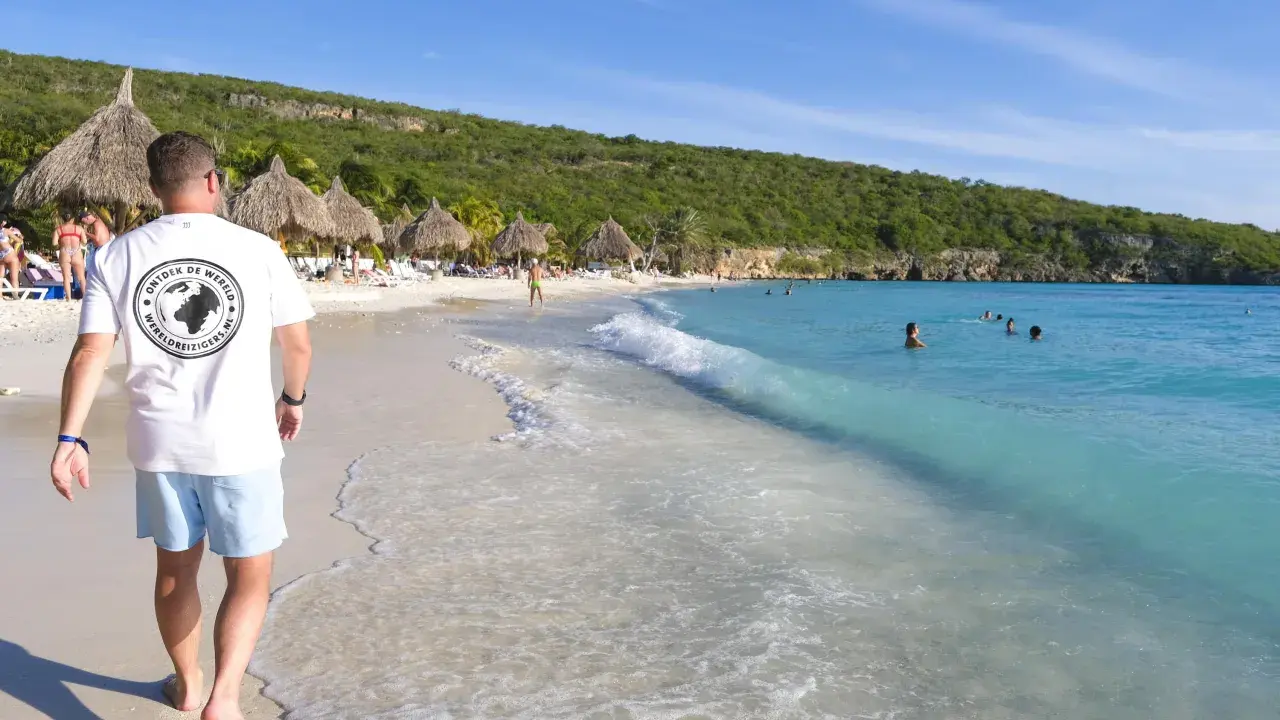 En man går längs en vit sandstrand med turkost vatten. Palmklippor och parasoll syns i bakgrunden. En perfekt plats att undra var ligger Curacao.