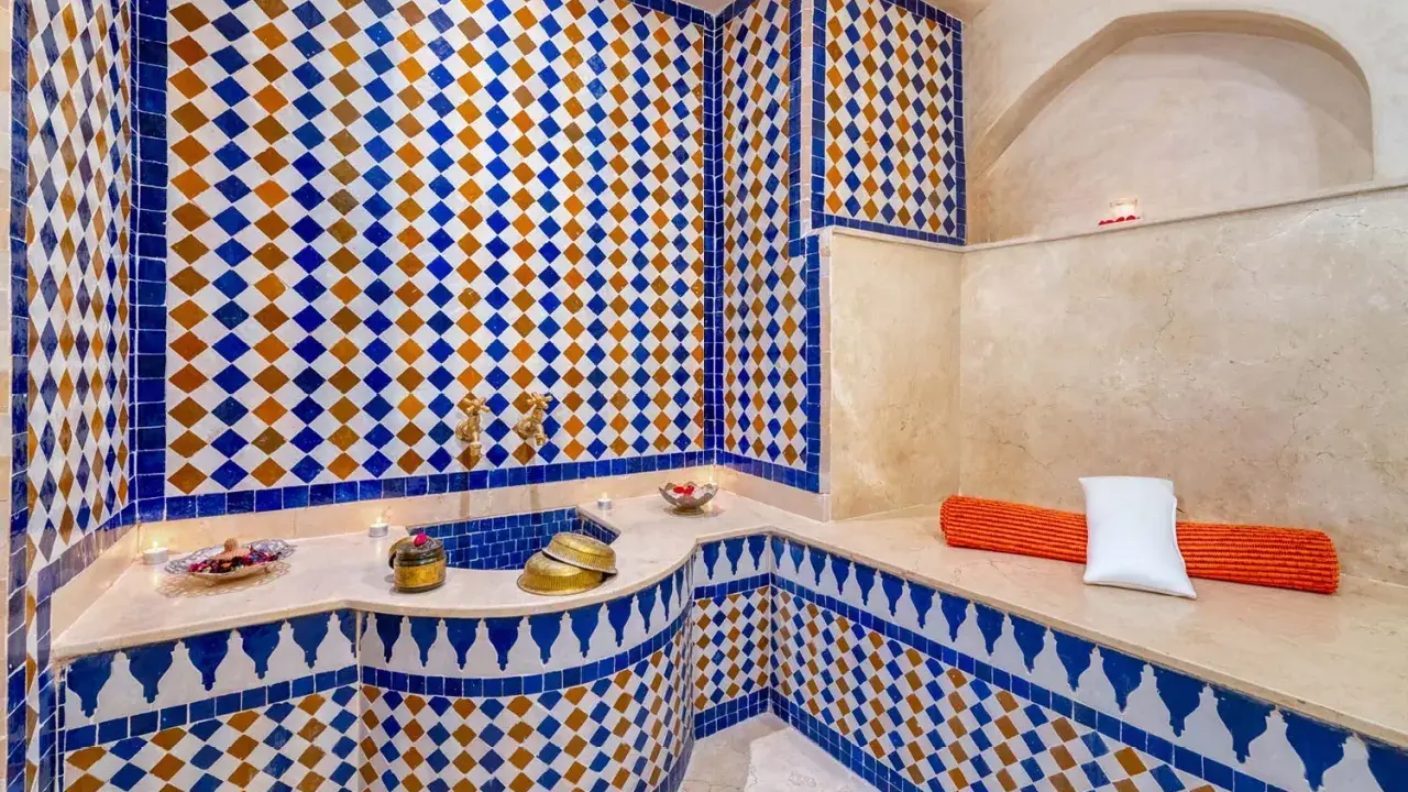 Ein luxuriöser Hamam in Marokko, verziert mit blauen und orangen Zellige-Fliesen. Kerzen und Schalen mit Rosenblättern schaffen eine entspannende Atmosphäre.