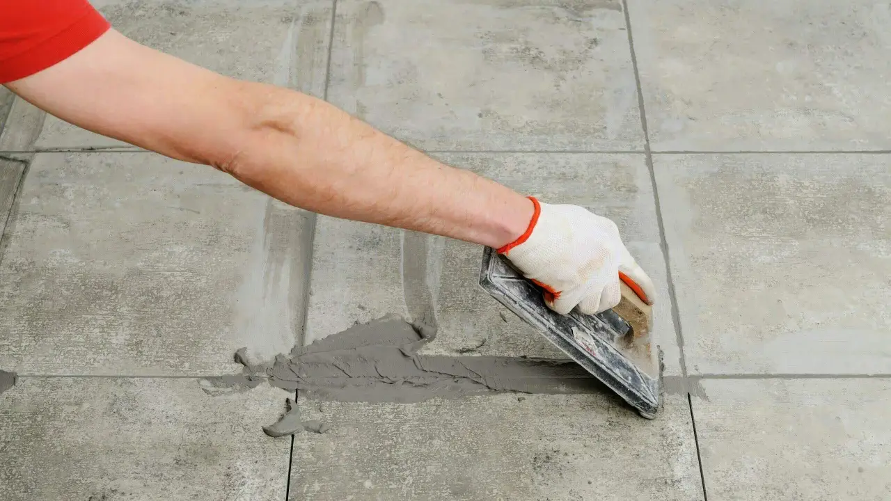 Mano con guante aplica lechada gris a las juntas entre azulejos de cemento con una llana.