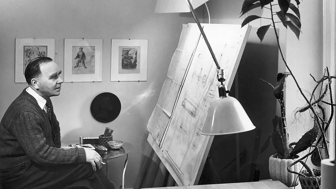 Paavo Tynell sitter vid sitt ritbord, omgiven av sina skisser och en stor ritning.
