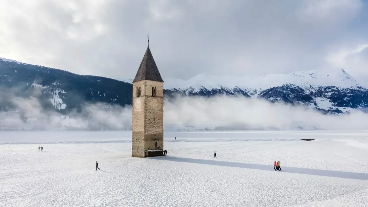 Torre sommersa nel lago di Resia, circondata da neve e montagne nebbiose. Alcune persone passeggiano sul ghiaccio.