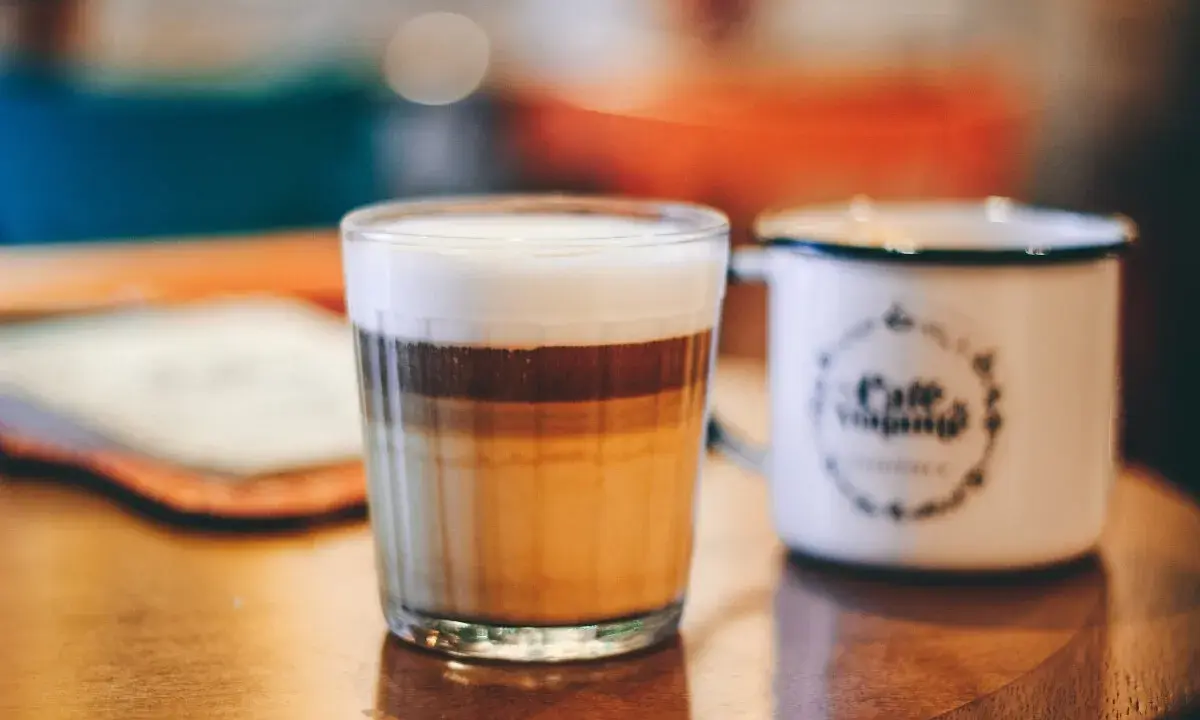 Ein geschichteter Latte Macchiato im Glas, daneben eine weiße Tasse mit Kaffee-Aufdruck.