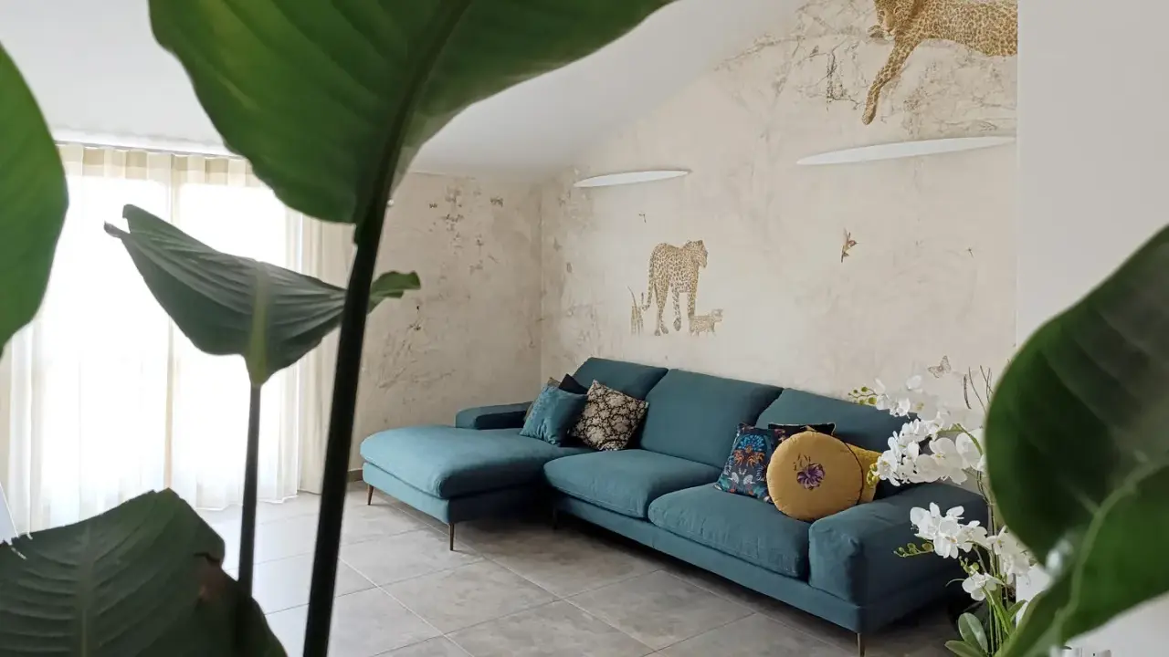 Divano turchese in un salotto con carta da parati a tema giungla e piante rigogliose, per un perfetto **stile jungle chic**.