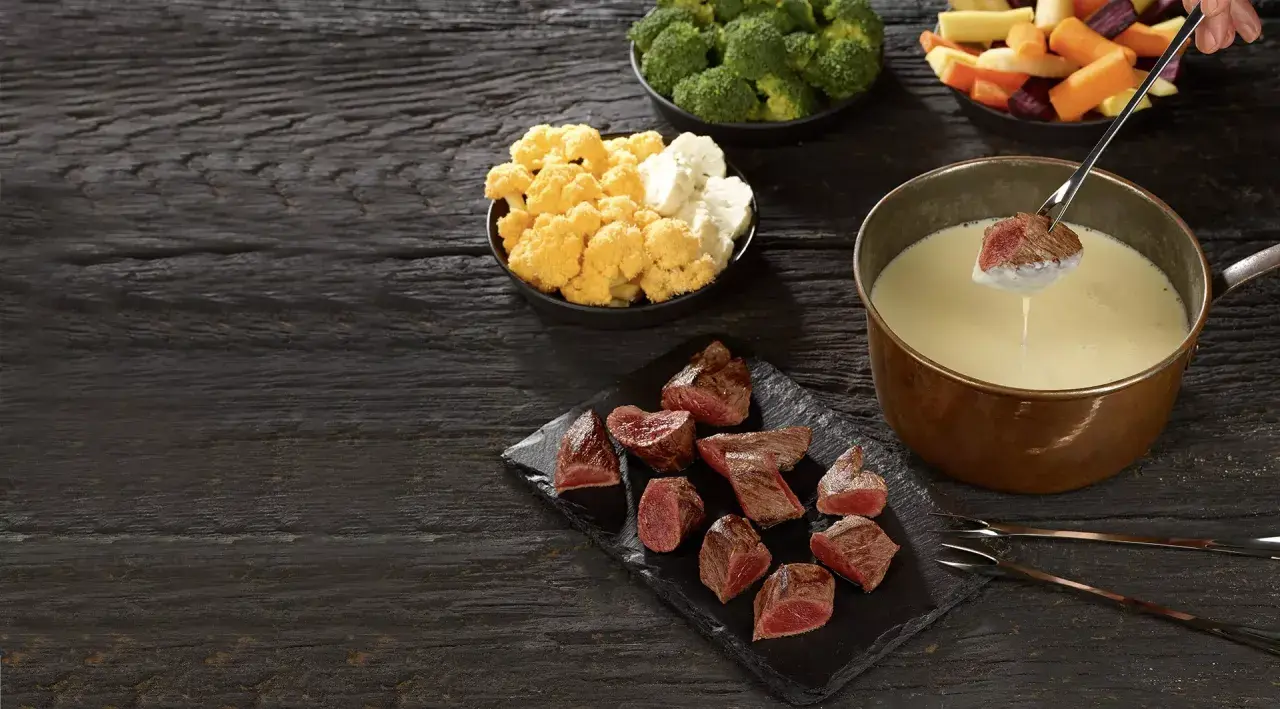 Un morceau de viande est trempé dans un caquelon de fondue. Des morceaux de viande, du brocoli, du chou-fleur et des légumes racines sont disposés sur une table en bois sombre.