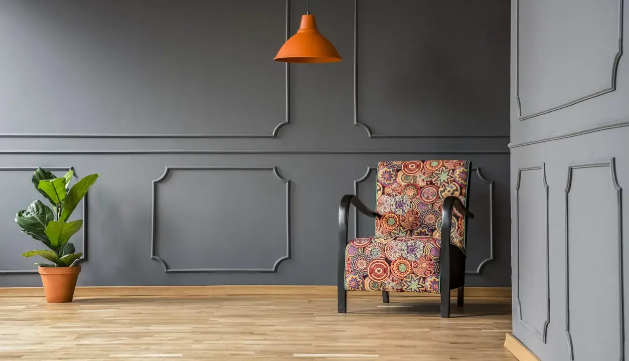 Nowoczesne sztukateria ścienna inspiracje: fotel w kolorowe wzory, pomarańczowa lampa i roślina na tle szarej ściany.