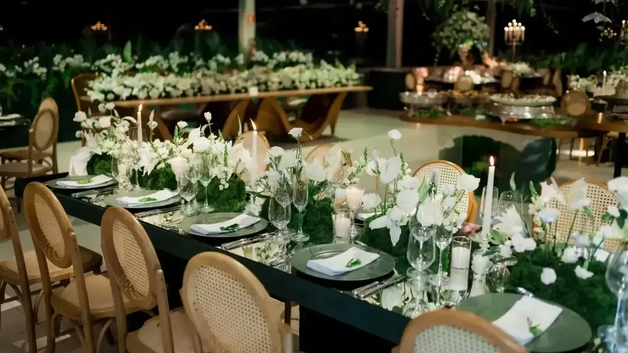 Eleganckie wesele na 100 osób. Długi, lustrzany stół z białymi kwiatami i świecami, otoczony wiklinowymi krzesłami.