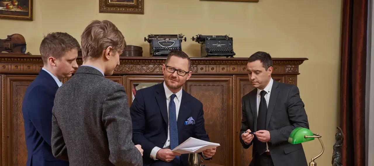 Czterech mężczyzn w garniturach rozmawia w gabinecie z zabytkowymi maszynami do pisania.