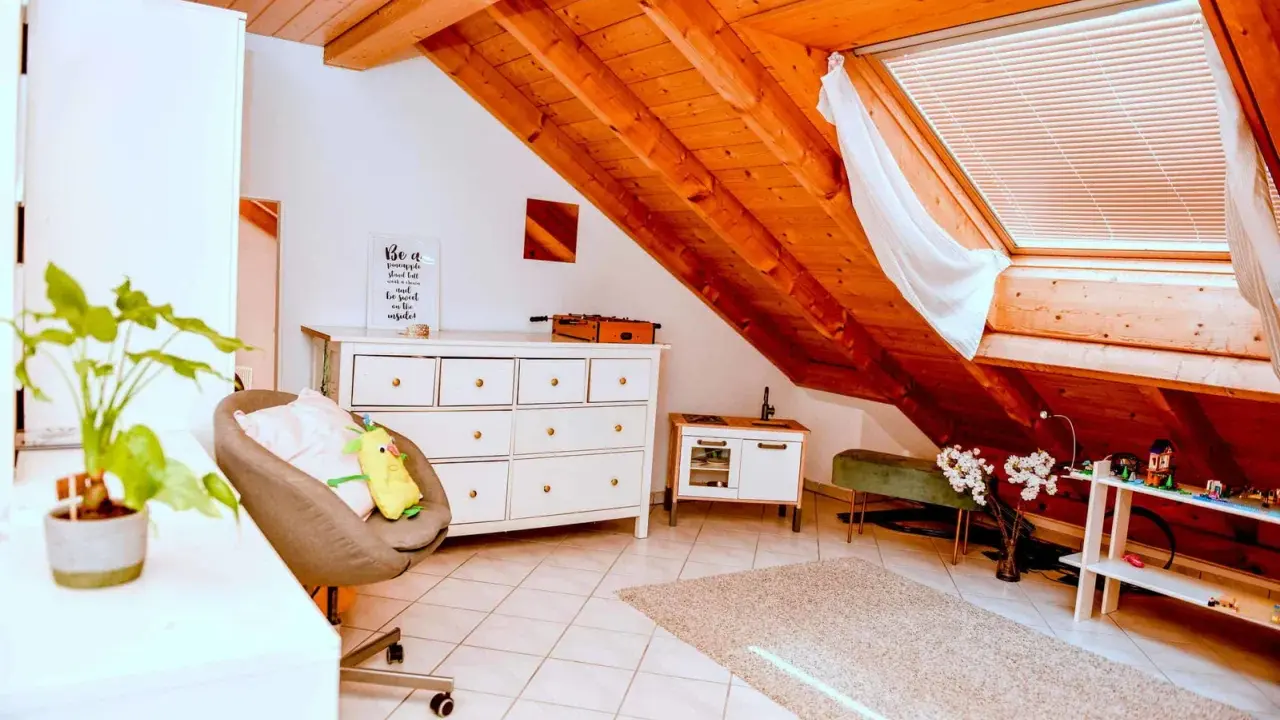 Acogedor despacho en buhardilla con vigas de madera, cómoda blanca, sillón y alfombra.