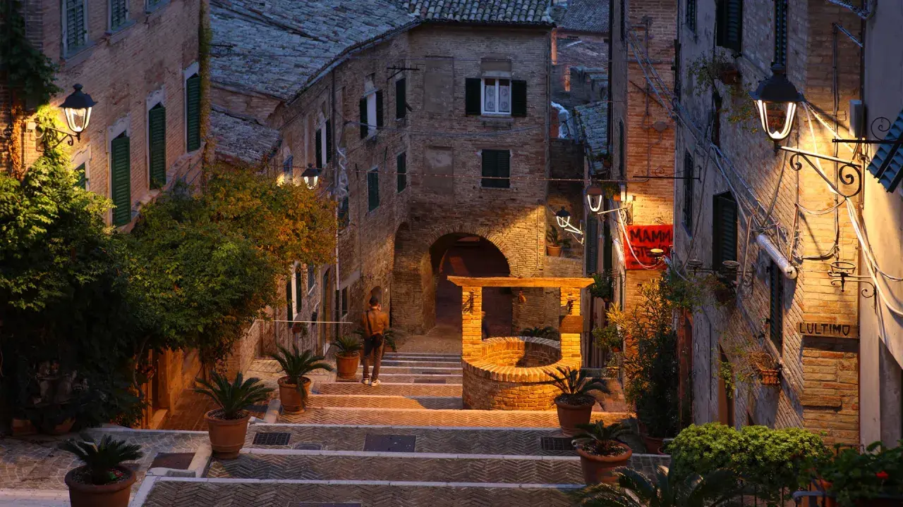 Antica scala a Gabicce Monte illuminata da lampioni, con un pozzo in mattoni e piante in vaso.
