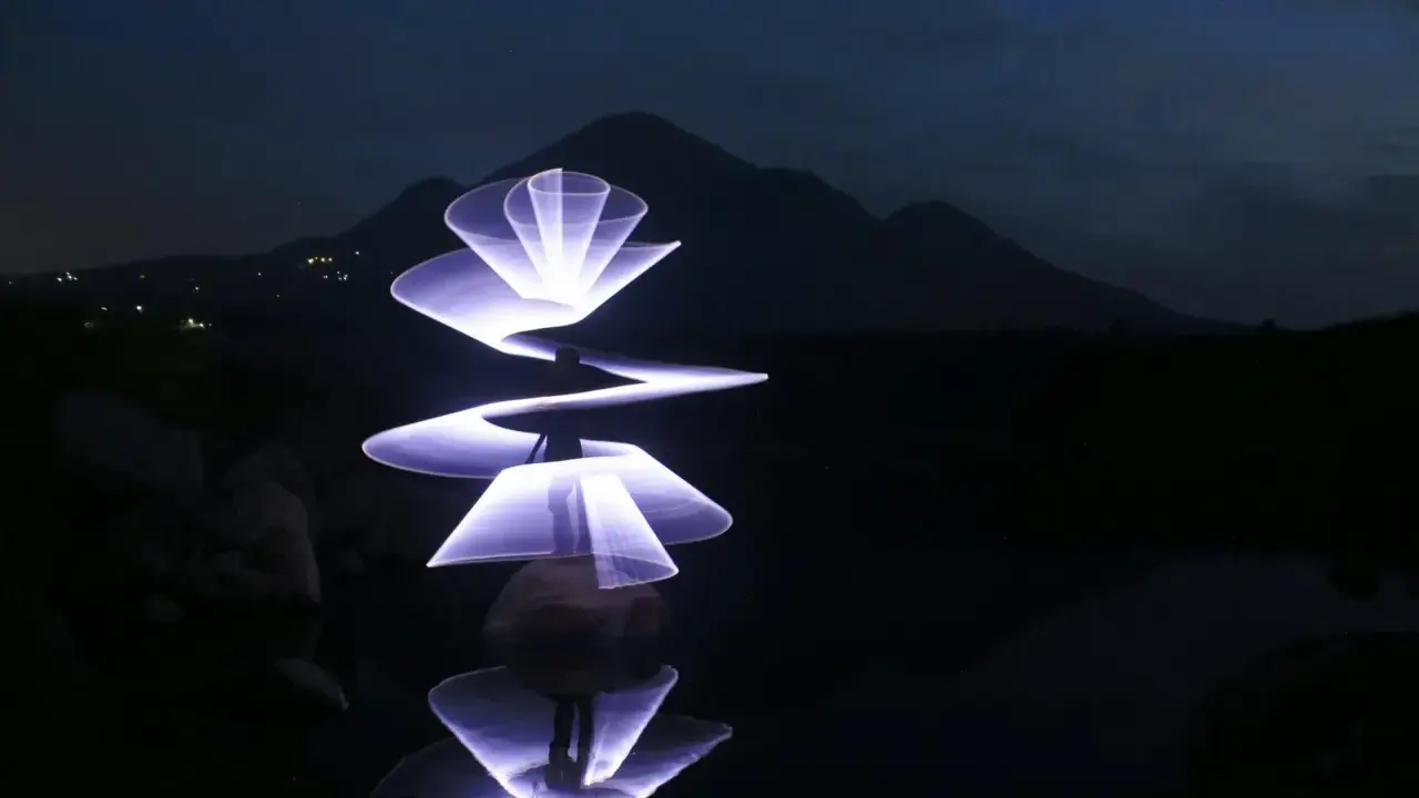 Un artista crea forme luminose con la light painting fotografia, disegnando spirali e petali di luce viola su uno sfondo scuro con montagne e riflessi sull'acqua.