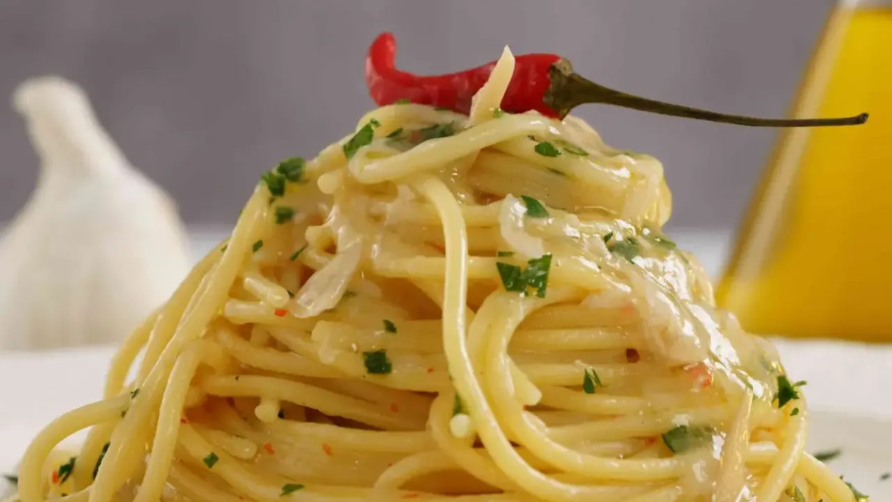 Spaghetti conditi con aglio, olio e peperoncino, guarniti con un peperoncino rosso e prezzemolo. Sullo sfondo, un bulbo d'aglio e una bottiglia d'olio.