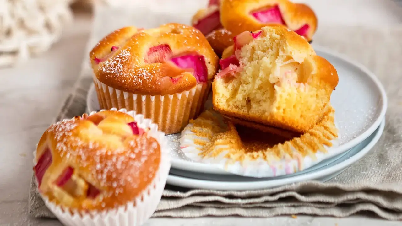 Saftige Rhabarber Muffins, mit Puderzucker bestäubt, liegen auf einem Teller. Einer ist angeschnitten und zeigt die rosa Rhabarberstücke.