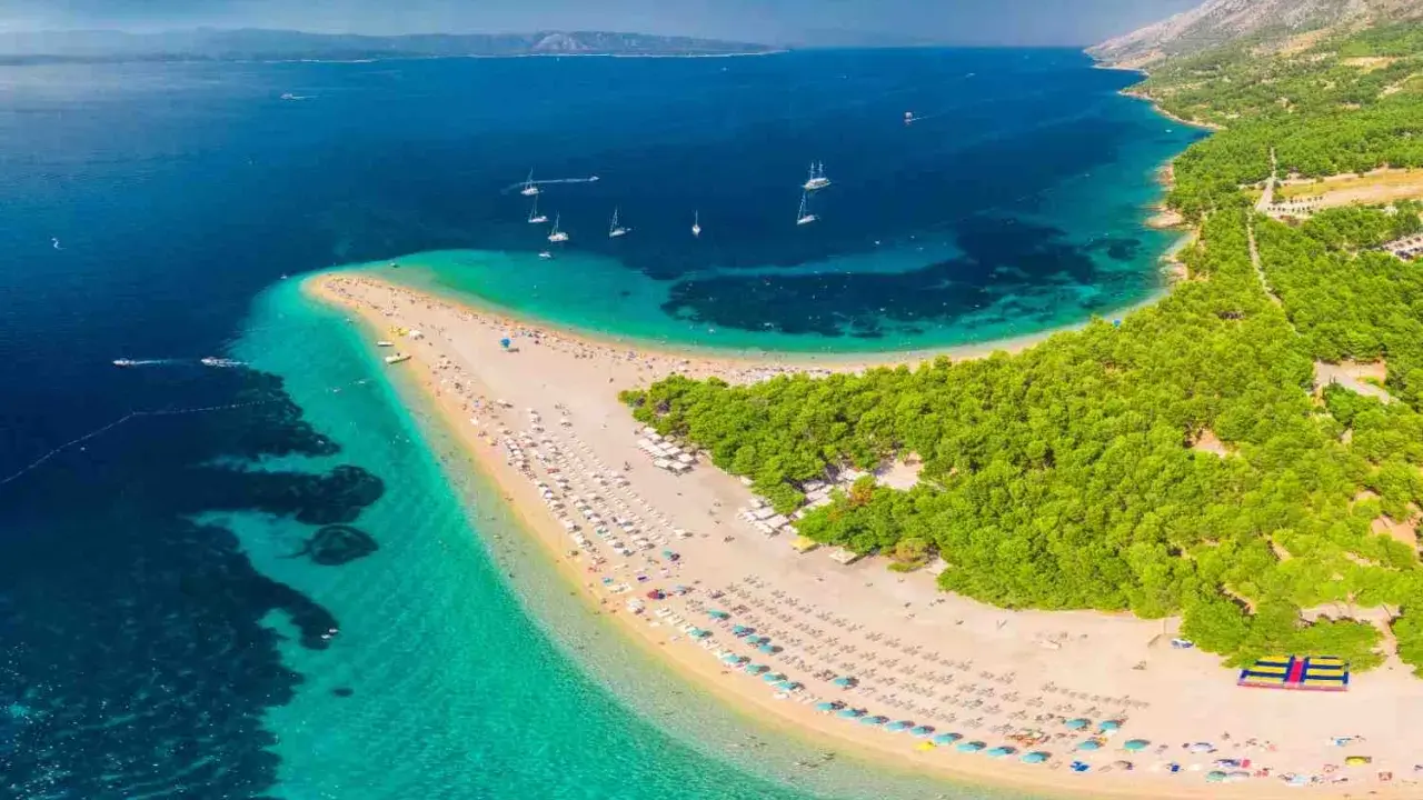 Plaża w kształcie języka, otoczona turkusową wodą i zielonym lasem, z widokiem na góry.