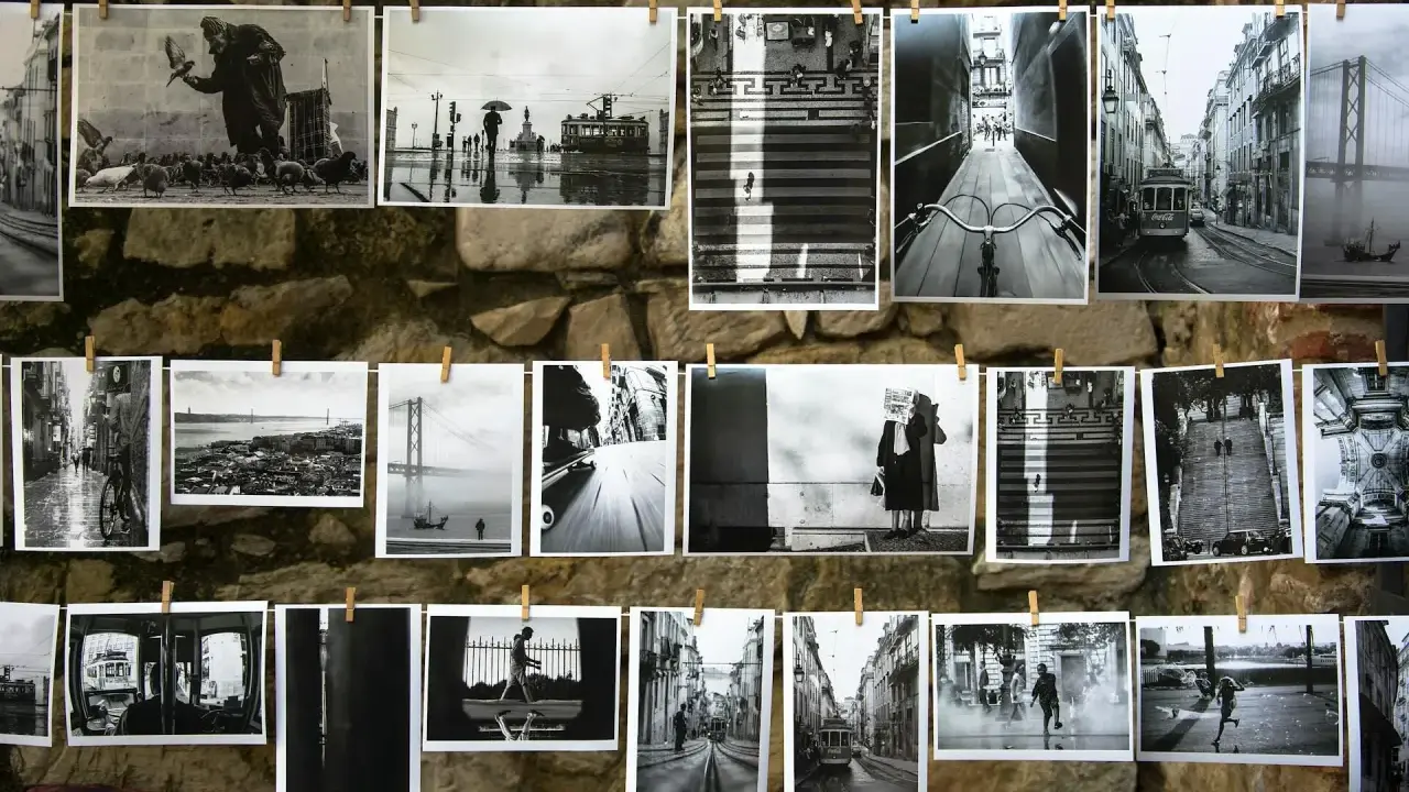 Czarno-białe fotografie wiszą na sznurku na kamiennej ścianie. Widać na nich uliczne sceny z Lizbony, w tym tramwaje, most i ludzi.