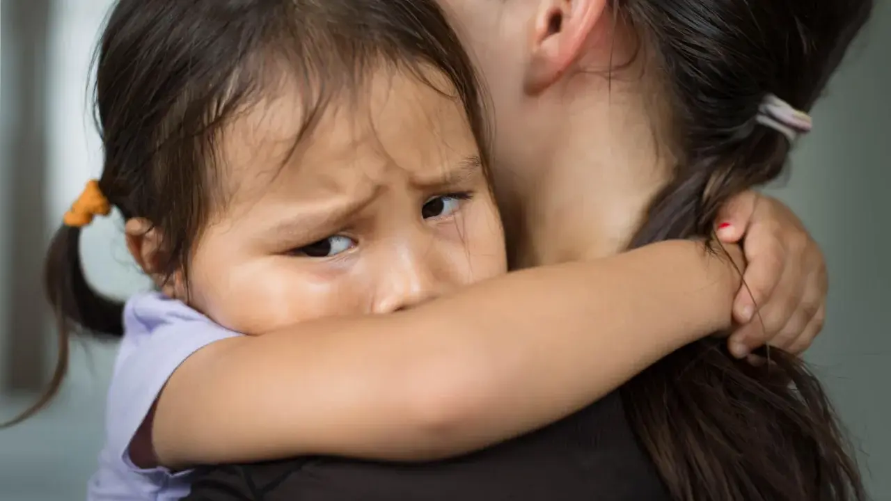 Ein Kind weint im Kindergarten und klammert sich an seine Mutter. Die Kleine hat Tränen in den Augen und sucht Trost.