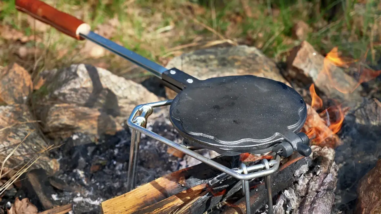 En gjutjärnspanna med trähandtag står på en ställning över en öppen eld. Perfekt för att steka plättar eller våfflor ute i naturen.