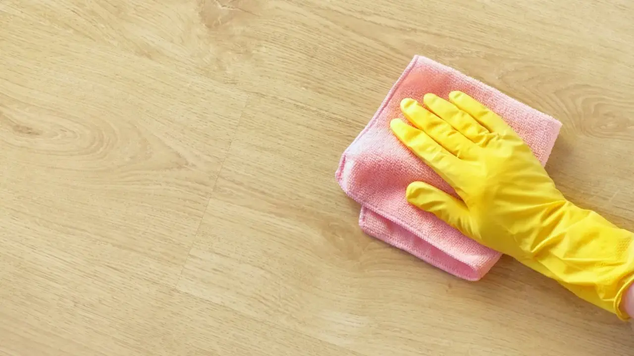 En hand i gul handske torkar av ett ljust trägolv med en rosa trasa. Perfekt för pigmenterad olja till ekbord.
