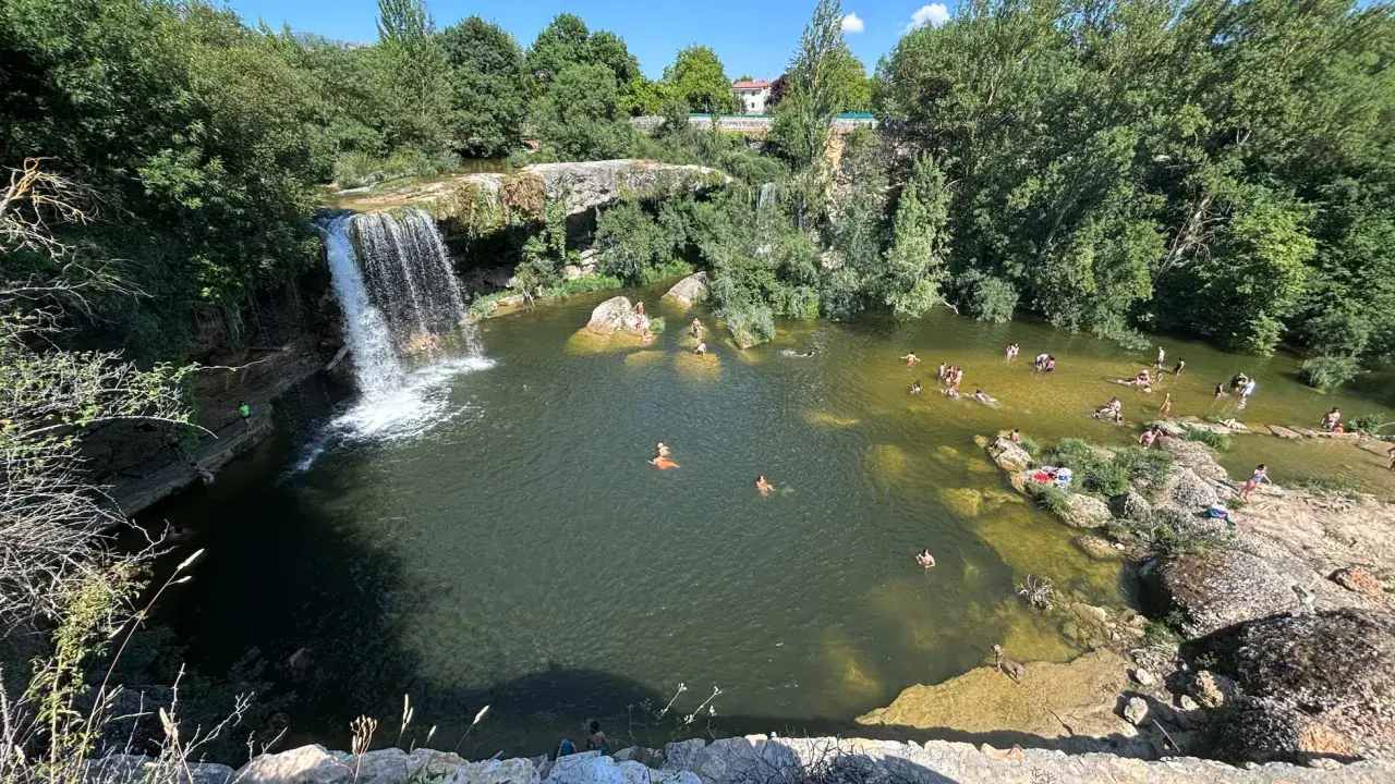 Gente disfrutando de un día soleado en las pozas del río Tus, con una cascada y vegetación exuberante.
