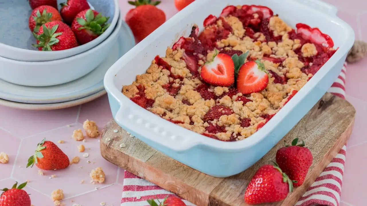 Ein frisch gebackener Erdbeer Crumble in einer hellblauen Auflaufform, garniert mit Erdbeerhälften. Daneben eine Schüssel voller Erdbeeren.