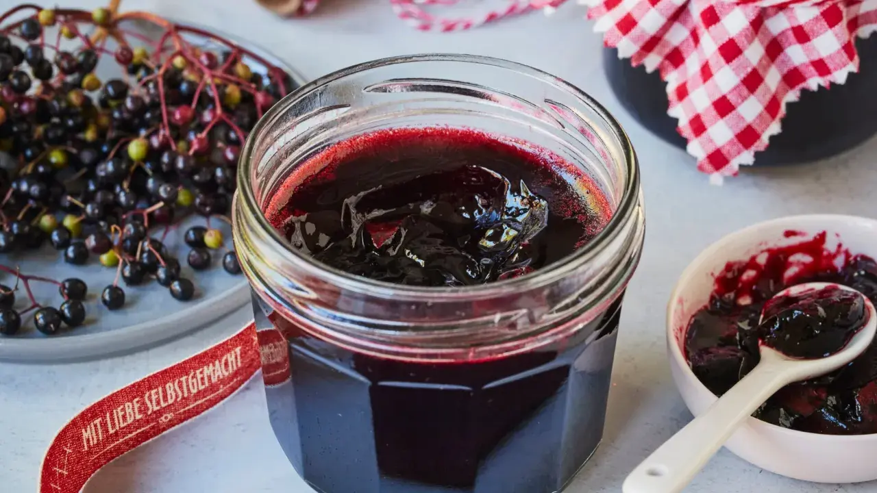Frisch zubereitete holunderbeeren marmelade im Glas, daneben frische Beeren und eine kleine Schale mit Löffel.