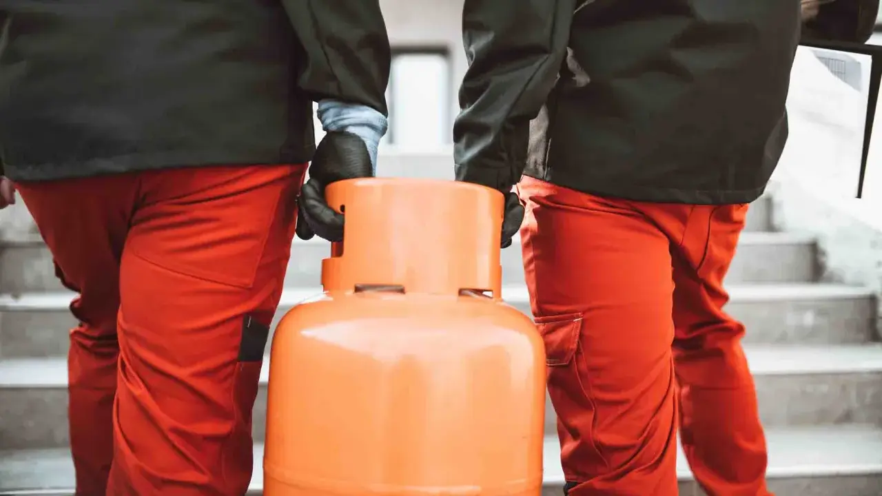 Dwóch mężczyzn w pomarańczowych spodniach niesie butlę z gazem. Wygląda na to, że nie można zakręcić butli z gazem, co może być problemem.