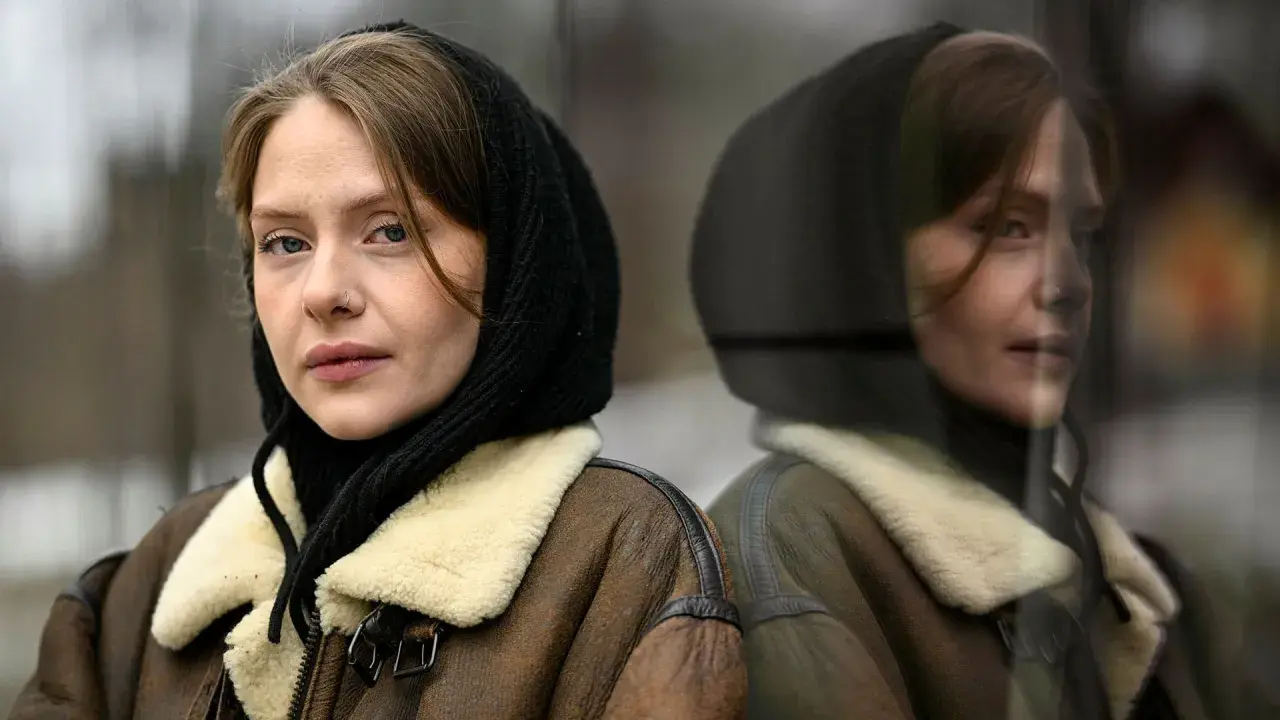 Sofia Kappel i en brun fårskinnsjacka med svart luva, blickar ut. Hennes spegelbild syns i en reflekterande yta.