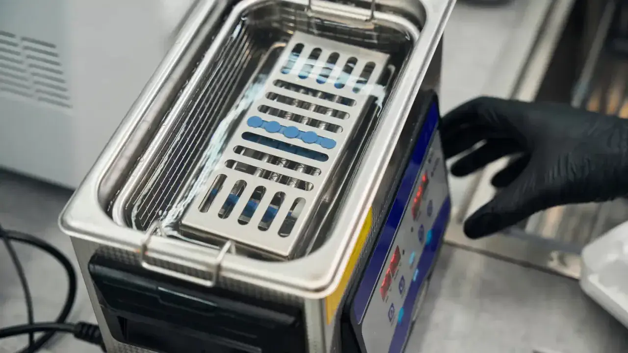 Ein Handschuh bedient ein Gerät, das zeigt, wie funktioniert ein ultraschallreiniger. Kleine Instrumente liegen in einem Korb im Wasserbad.