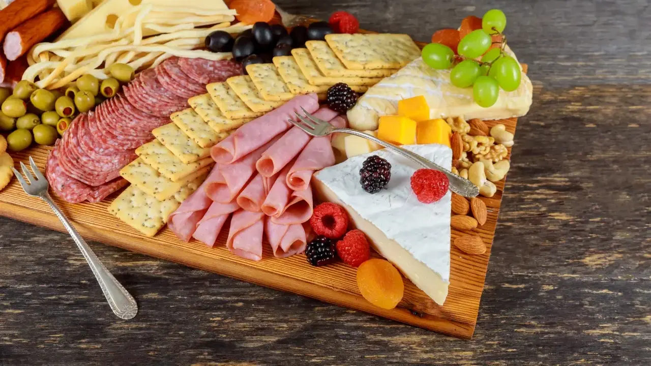 Apetyczna deska serów i wędlin: salami, szynka, ser żółty, pleśniowy, oliwki, winogrona, maliny, orzechy i krakersy.