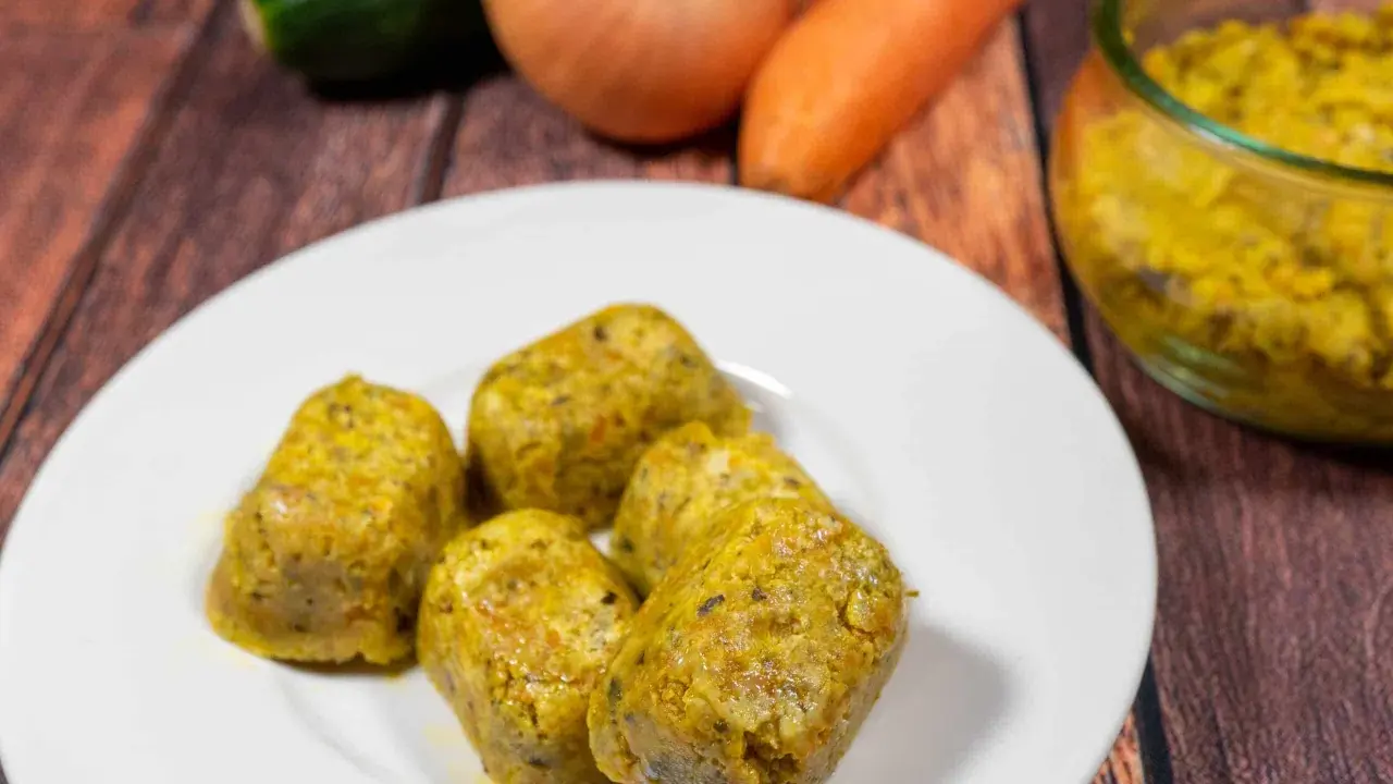 Cinque cubetti dorati di dado vegetale fatto in casa, preparati con carote, zucchine e cipolle, su un piatto bianco.