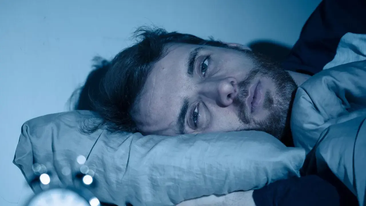 Mann liegt wach im Bett, Augen weit offen, die Angst vor Schlaflosigkeit ist ihm ins Gesicht geschrieben. Ein Wecker ist im Vordergrund zu sehen.