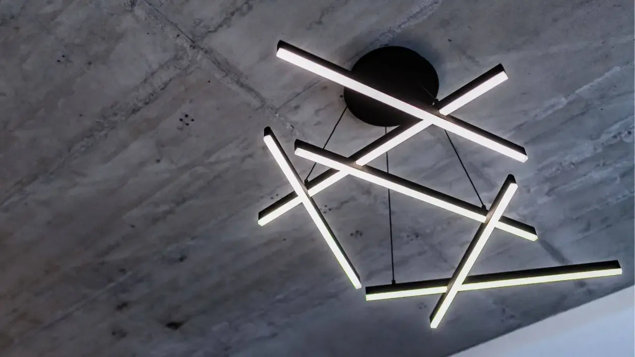 Oryginalna lampa sufitowa z geometrycznymi, podświetlanymi ramionami, zawieszona na tle surowego, betonowego stropu.