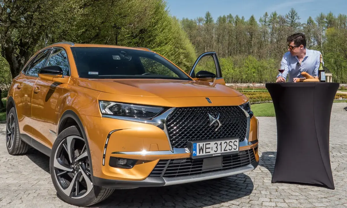 Pomarańczowy Citroën DS7 Crossback z otwartymi drzwiami, obok stolika z winem i bagietką, na tle zieleni.