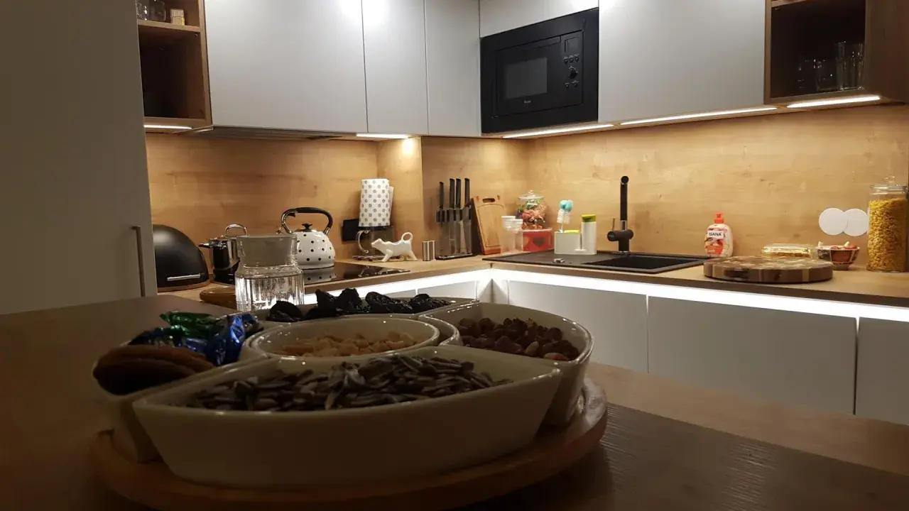 Nowoczesna kuchnia z białymi frontami i drewnianym blatem. Na blacie czajnik, zlew i naczynia z bakaliami. W tle wbudowany czarny okap, który idealnie komponuje się z całością, pokazując jak obudować komin w kuchni.