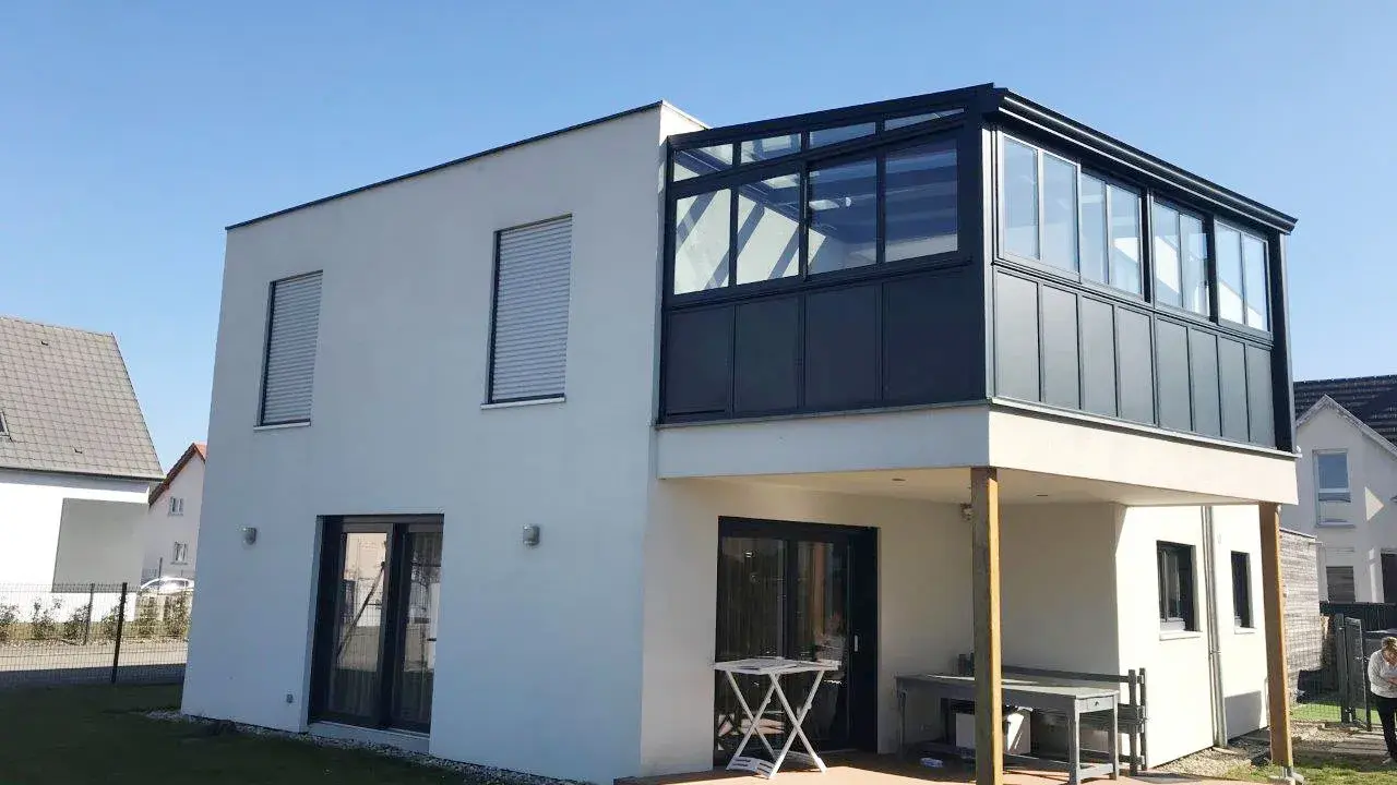 Maison moderne avec extension étage sur piloti. Une véranda vitrée surplombe une terrasse en bois.