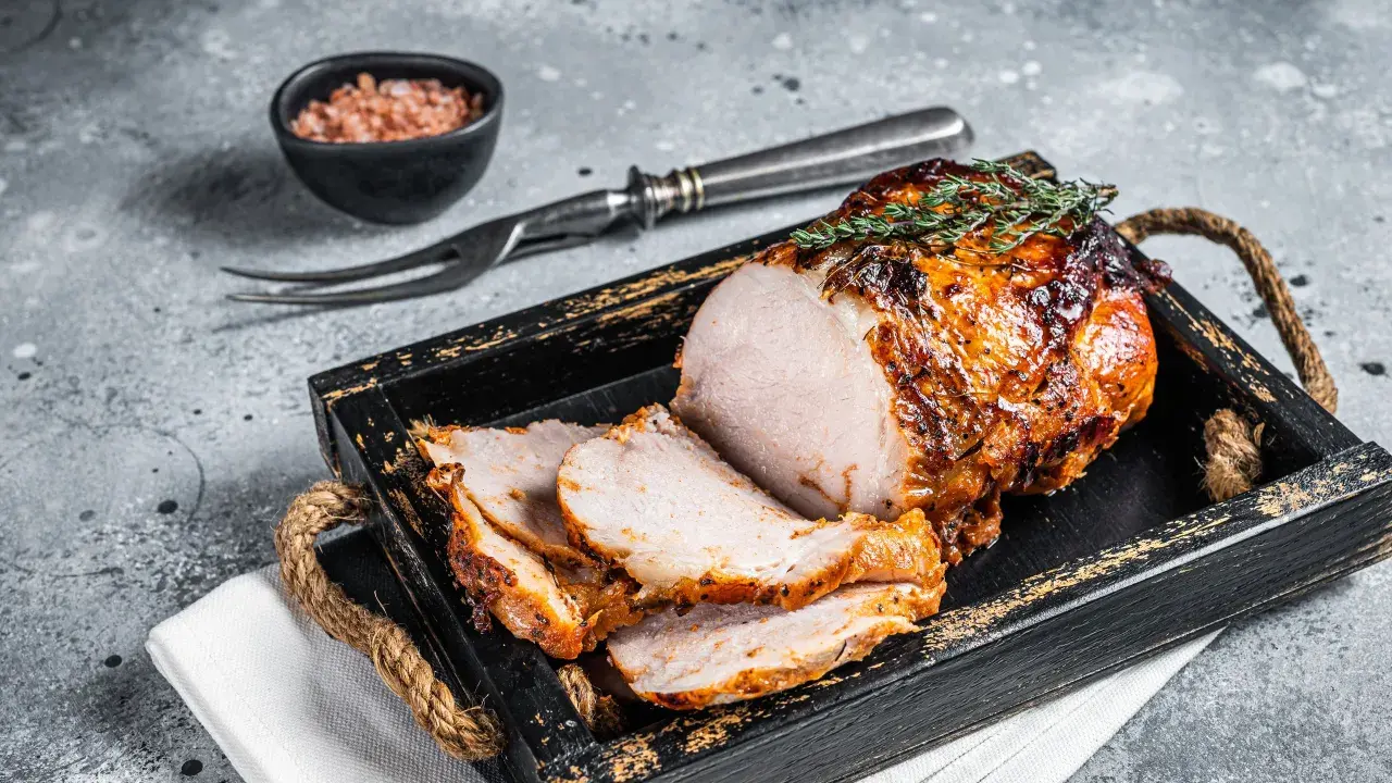A sliced, roasted pork loin with a golden-brown crust, garnished with thyme, on a rustic wooden tray.