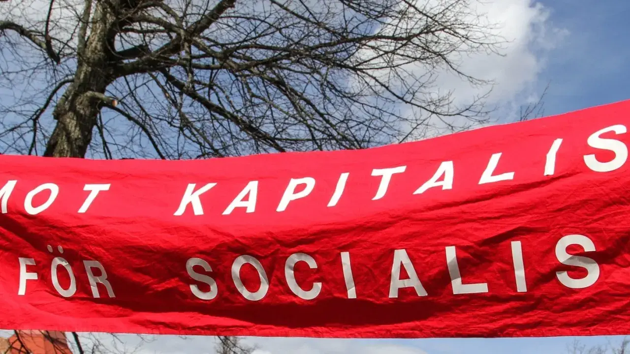 Röd banderoll med vit text: "MOT KAPITALIS FÖR SOCIALIS". En demonstration som visar vad socialism betyder.