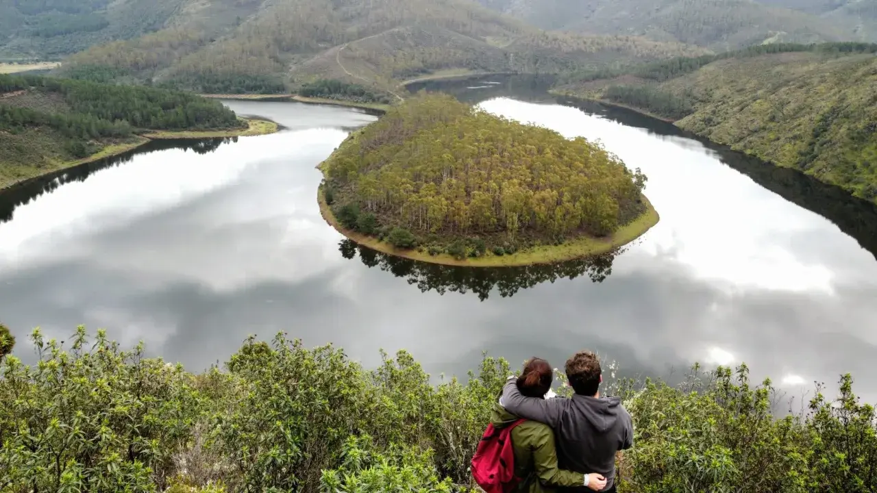 Pareja abraza contemplando el meandro del río en Las Hurdes, un paisaje de colinas verdes y agua serena.