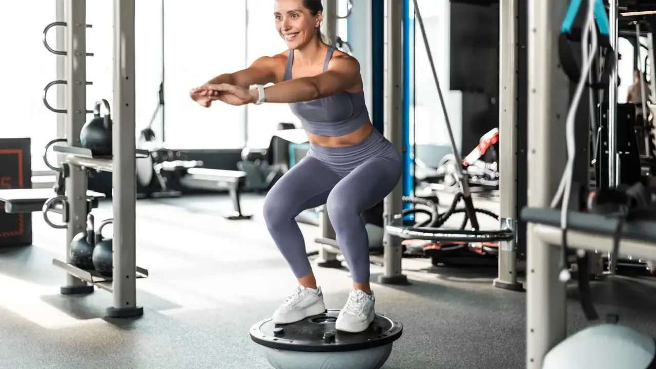 Frau macht Kniebeugen auf einem Bosu-Ball, trägt Sportkleidung und weiße Sneaker. Sie trainiert ohne schuhe auf dem instabilen Untergrund.