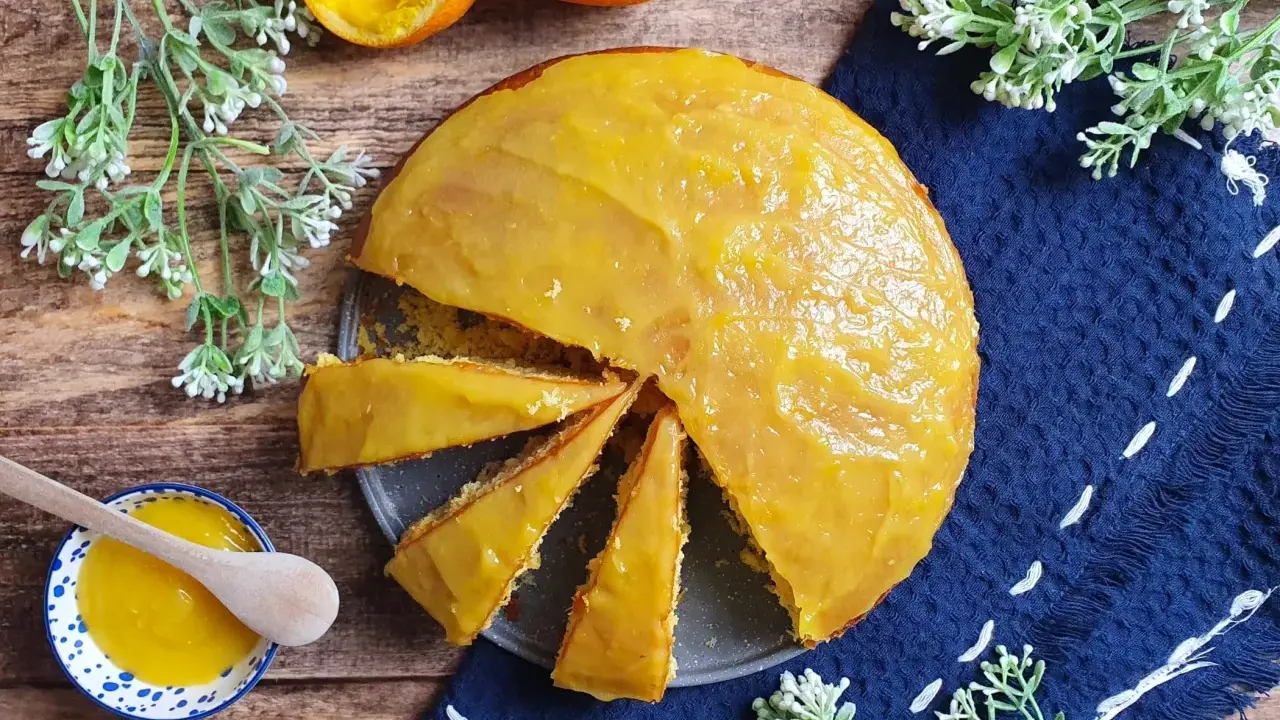 Una torta all'arancia morbidissima, con glassa lucida e profumata, pronta per essere gustata. Accanto, una ciotolina con crema all'arancia e un cucchiaino di legno.
