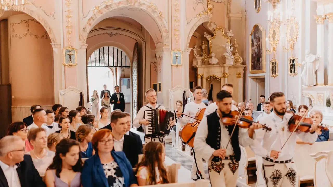 Muzycy w strojach ludowych grają tradycyjne pieśni na ślub w kościele. W tle para młoda wchodzi do świątyni.