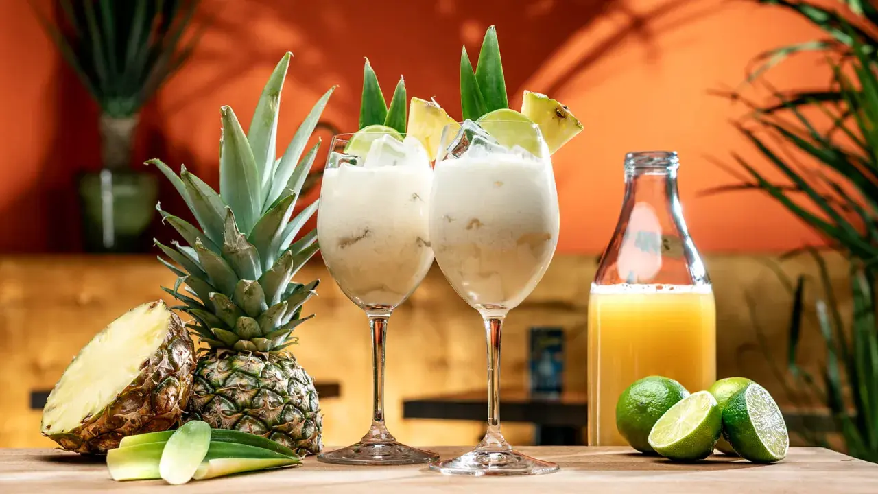 Två krämiga drinkar med ananasjuice, is och lime. En hel och en halv ananas samt en flaska juice står bredvid.