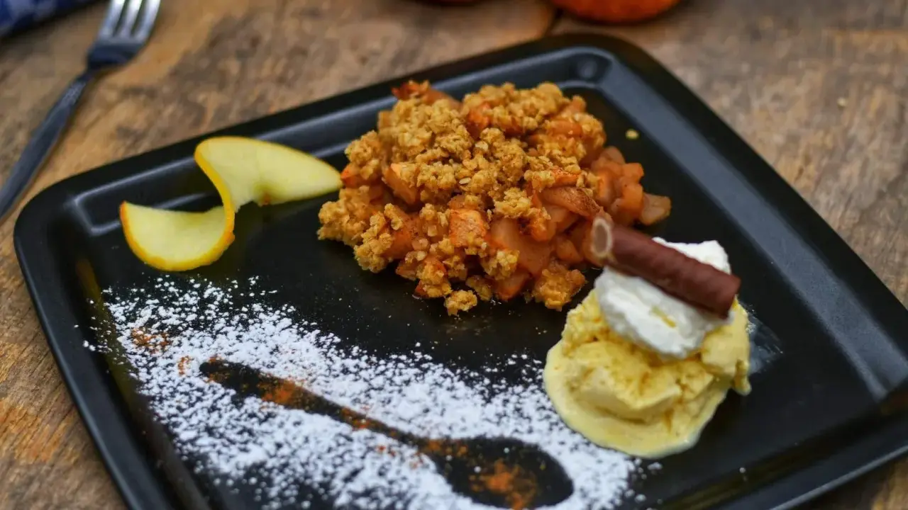 Ein köstlicher Apfel Crumble mit Haferflocken, serviert mit Vanilleeis und Sahne. Ein süßer Genuss auf einem schwarzen Teller, verziert mit Puderzucker.