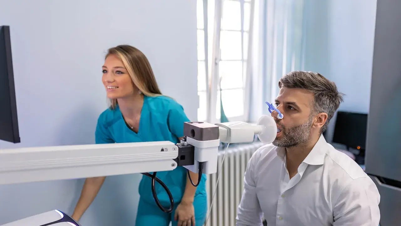 Mann macht Lungenfunktionstest, Arztin überwacht. Ergebnisse zeigen 70 Prozent Lungenfunktion.