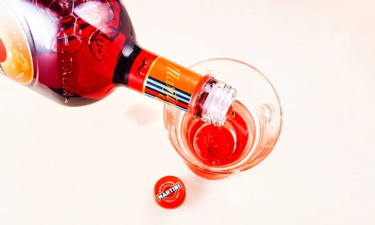 Czerwony Martini Fierro nalewany do kieliszka. Obok leży czerwona zakrętka z logo Martini.