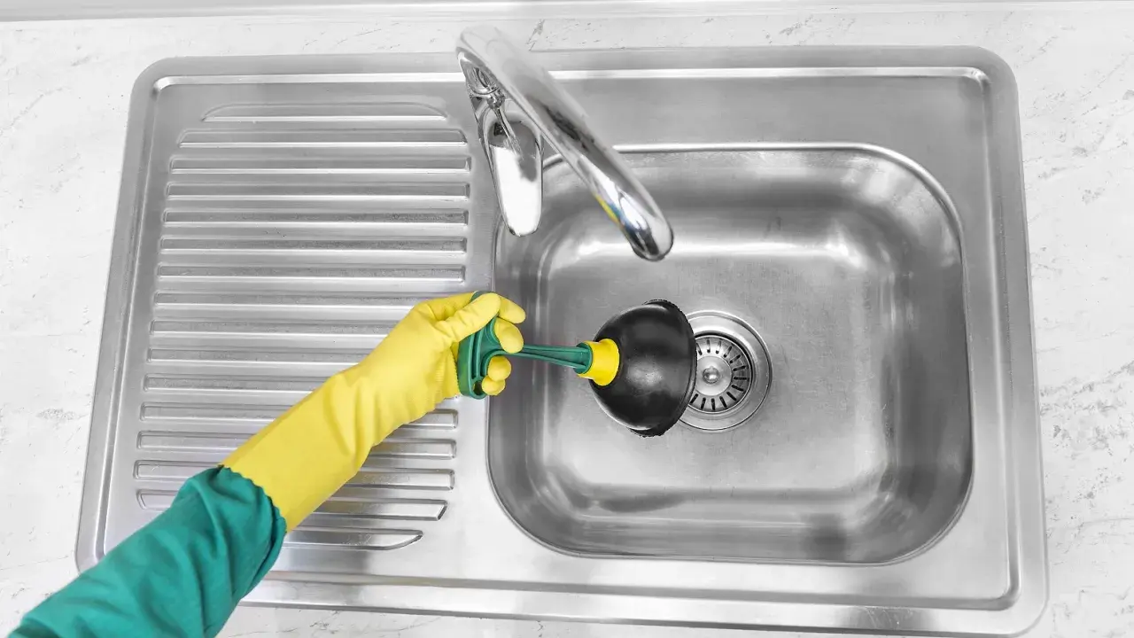 Mano con guante amarillo usando desatascador para limpiar tuberías del fregadero.