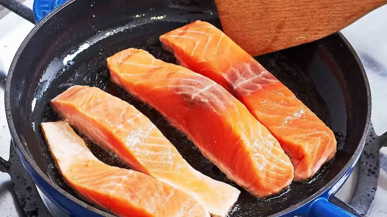 Vier Lachsfilets braten in einer Pfanne. Ein Holzspatel liegt bereit, um zu zeigen, wie bereite ich Lachs zu.