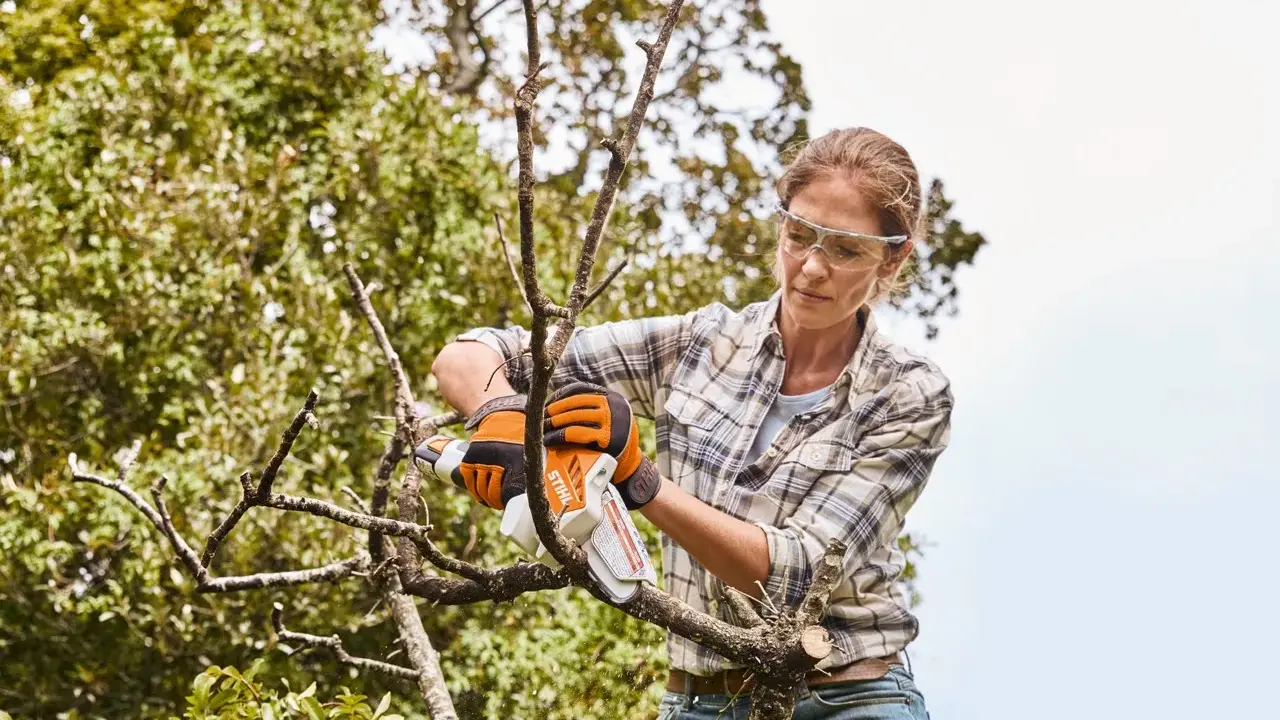 Femme en lunettes de protection et gants orange, effectuant l'entretien des arbres avec une tronçonneuse.