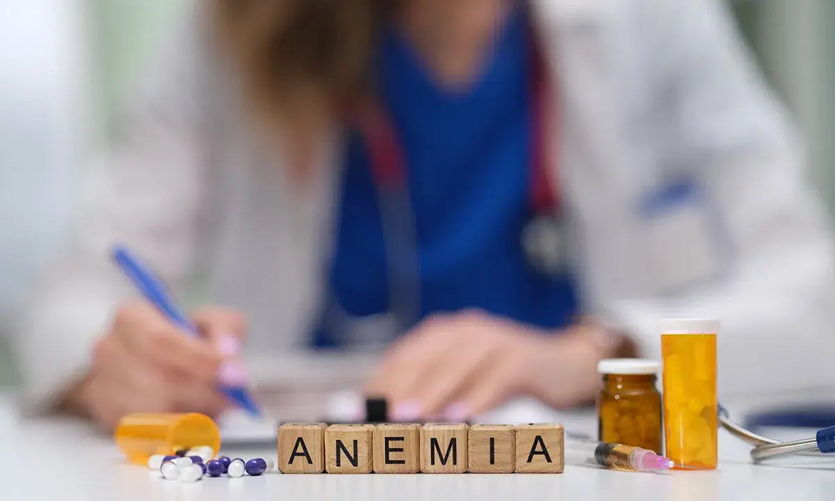 Lekarz zapisuje zalecenia dotyczące leczenia anemii u starszej osoby. Na stole leki i kostki z napisem ANEMIA.