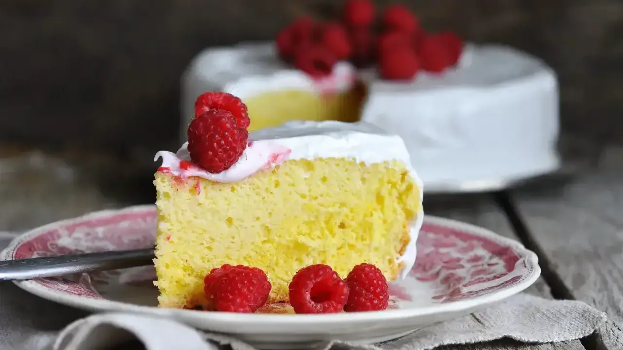 Wilgotne ciasto tres leches z malinami na talerzyku, w tle cały tort.