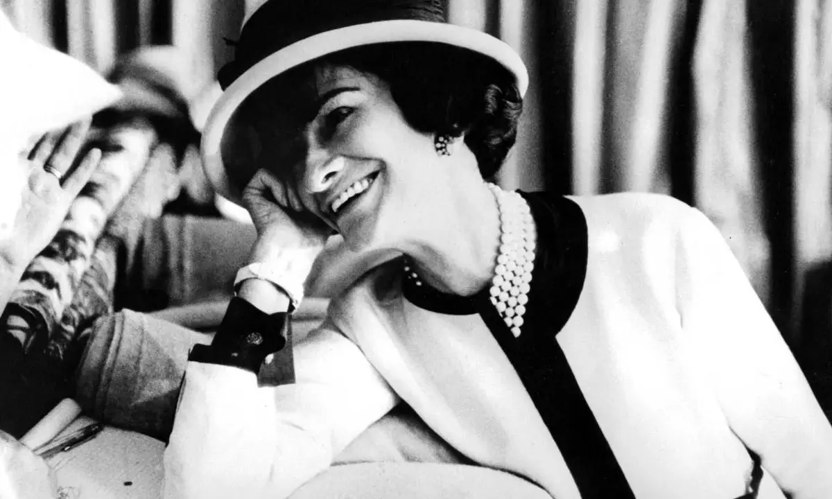 Coco Chanel, sonriendo con un sombrero y perlas, evoca la elegancia de donde es Chanel.